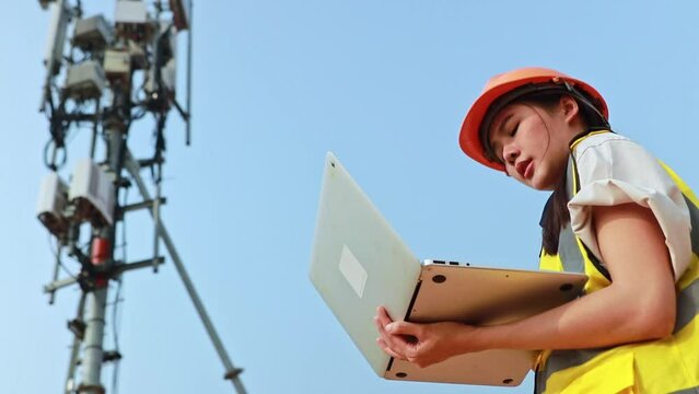 Asian female engineer wearing helmet performs field work telecommunications tower controlling mobile power installation using laptop maintain 5G network installed high rise building.