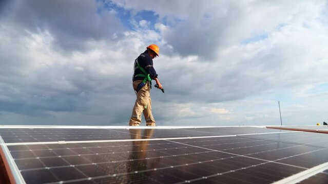 Asian male electrician engineer inspecting installation solar panels on metal roof industrial factory walking around holding power drill check important points each solar panel on the clipboard.