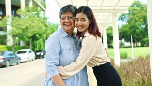 Family portrait happy elderly mother and her cute beautiful healthy asian daughter stand in an embrace smiling and good mood looking at the camera while standing in an outdoor park.