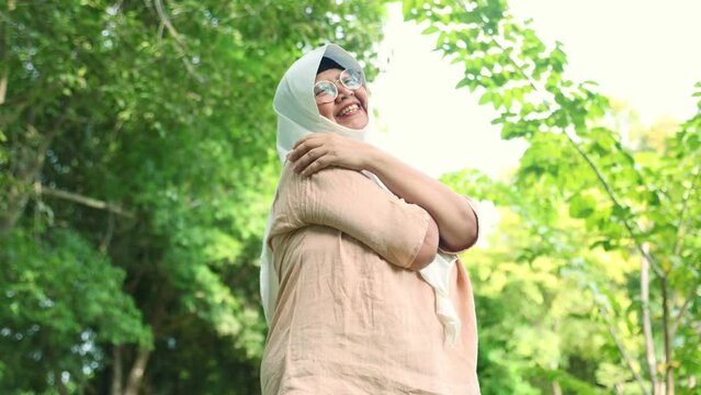 Elderly asian muslim woman wearing hijab positive emotions standing and embracing the cool breeze beautiful nature green trees shady trees good weather in the garden makes for good mental health.