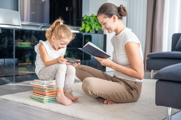 Little girl sits on a stack of children's books and uses her smartphone while her mother reads a...