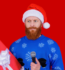 Santa Claus bearded man in Xmas hat and Christmas winter sweater hold present and microphone for...