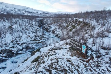 The drone hovers above Björkliden's trailhead sign, showcasing a meandering frozen creek through a sparse snowy forest