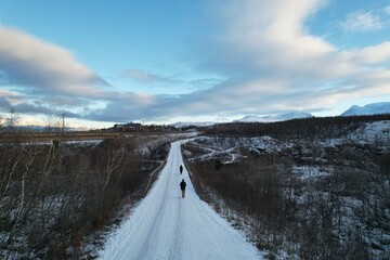 An aerial view of a remote Björkliden trail reveals a snow-laden path flanked by bare trees and a solitary walker amidst the vastness.