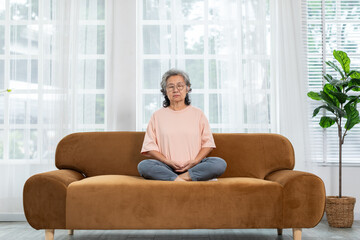 Mature Asian woman doing yoga for meditation on sofa in living room at home.Clam of senior or...