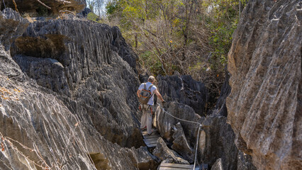 A boardwalk with safety cables runs between the steep karst rocks. A man with a backpack walks along the path, holding on to the cliffs. Thickets of forest ahead. Madagascar. Tsingy De Bemaraha 