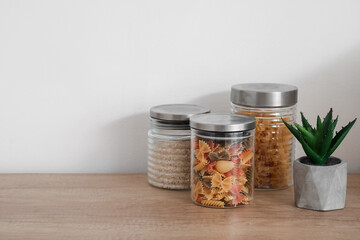 Jars with different types of pasta, rice and houseplant on white kitchen counter