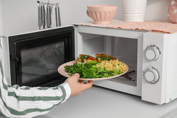 Woman putting plate with pasta into microwave oven in modern kitchen