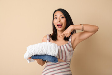 Beautiful young woman pointing at stack of clean clothes on beige background