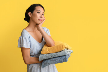 Thoughtful young woman with stack of clean clothes on yellow background