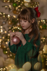 A portrait of a sweet beautiful smiling little 7-old girl wearing green dress, with red bow on the her brown long hair, decorate a Christmas tree with a pink shine glass ball at home at Christmas eve