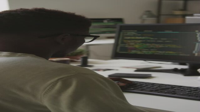 Vertical rear view waist up shot of African American guy in t-shirt and eyeglasses developing new software on desktop computer in office, writing programming codes via keyboard