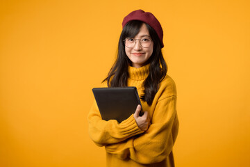 Standing out in a yellow sweater and red beret, young Asian woman works on her laptop against a cheerful yellow backdrop.