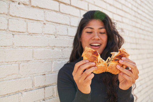Woman eating savory pastry outdoors in front of white brick wall pulling pastry apart