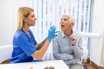 Senior patient opening her mouth for the doctor to look in her throat. Female doctor examining sore throat of patient in clinic. Otolaryngologist examines sore throat of patient.