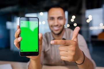 Smiling man holding a phone with a green screen in his hands, showing it to the camera and pointing at it with his index finger, looking at the camera. - Powered by Adobe