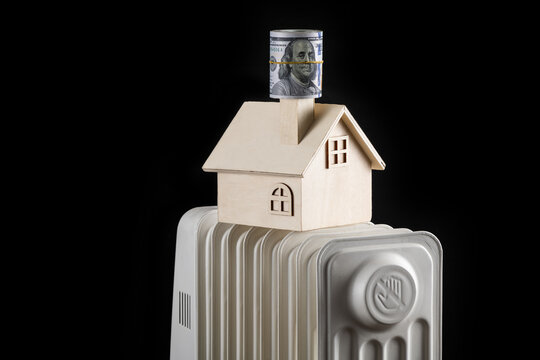 A Stack Of 100 American Dollars Banknotes, A Symbolic Wooden House And An Electric Heating Battery On A Black Background