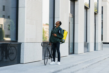 An African-American male courier with a backpack walks next to a bicycle and looks for the entrance to the building. Delivery from the restaurant