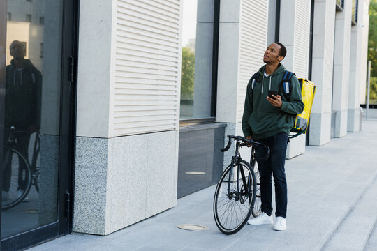 A dark-skinned male courier walks with a bicycle along the building and looks at the phone screen. The man looks at the address on the map