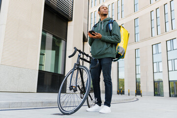 A bicycle courier with a yellow backpack holds a phone in his hands against the background of a...