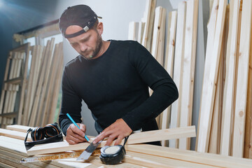 A carpenter man in dark clothes marks future holes in the board with a pencil. Manufacture of furniture made of wood in your workshop. - Powered by Adobe