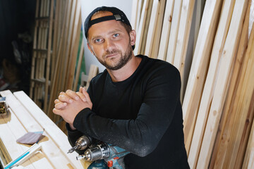 A successful male carpenter with a beard in dark clothes stands against the background of his workshop.