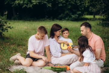 Fototapeta premium Big family sitting on the picnic blanket in city park during weekend Sunday sunny day. They are smiling, laughing and eating boiled corn and watermelon.