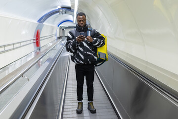 An African-American courier is walking in a subway crossing with a mobile phone. Yellow backpack on...