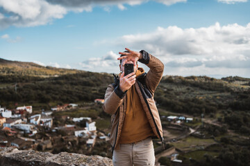 Young happy photographer man traveling sightseeing in a small town, taking a photo with his cell...