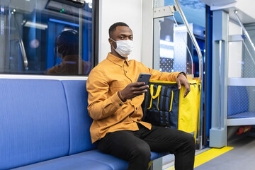 An African-American courier in a protective mask holds a mobile phone while riding in a subway car with a yellow backpack