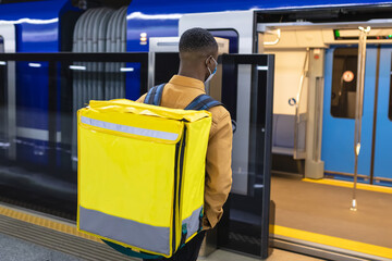 An unrecognisable deliveryman in a protective mask with a yellow backpack enters the subway car....
