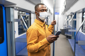 An African-American businessman in a protective mask with a mobile phone rides in a subway car