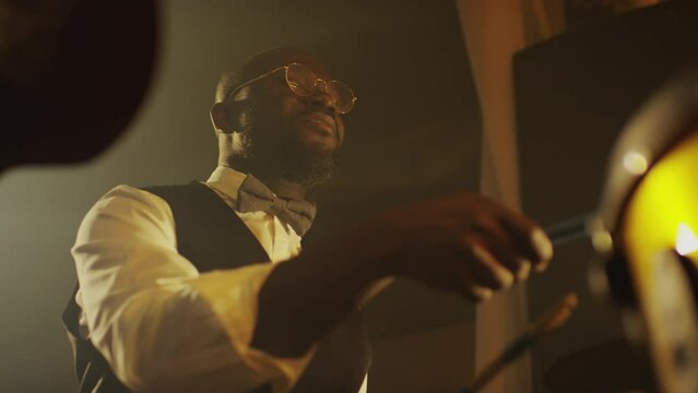 Low angle of Black male jazz musician wearing elegant vest and bow tie playing drums on smoky stage in night club