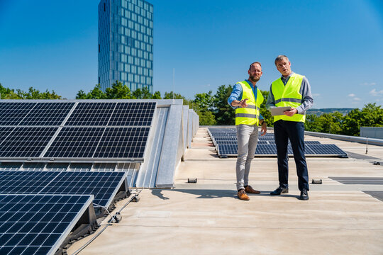 Two workers in safety vests discussing on the rooftop of a solar-powered company building while holding a tablet