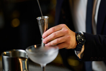 The hands of an unrecognisable bartender who pours a cocktail ingredient into a measuring glass....