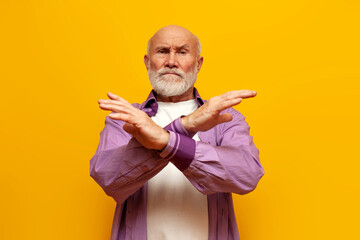 old bald grandfather in purple shirt prohibits and shows stop gesture on yellow isolated background