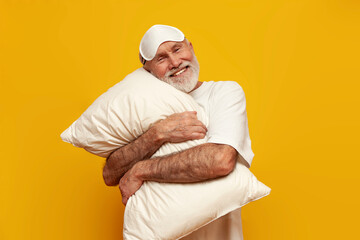 cheerful old bald grandfather in white t-shirt and sleep mask holds and hugs pillow on yellow...