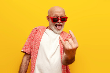 old grandfather pensioner in sunglasses shows rock gesture and sings to the music at party on yellow isolated background