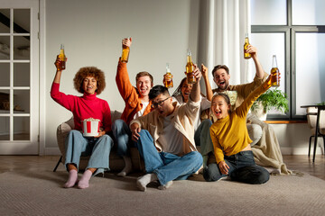 multiracial group of young friends sitting on the couch at home drinking beer and watching TV and celebrating good luck