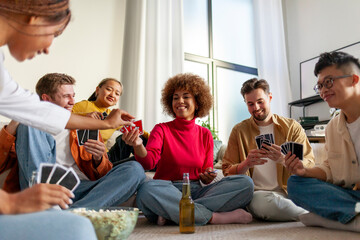 multiracial group of young friends playing cards drinking beer and having fun at home, students...
