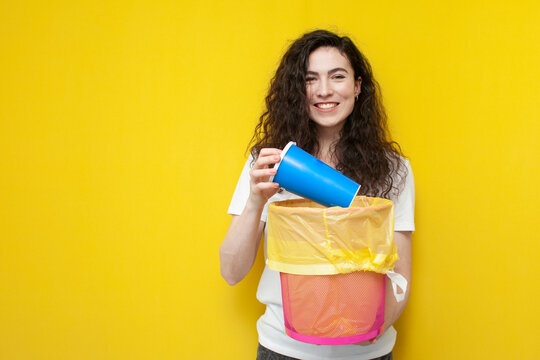 young cheerful girl throws plastic glass into a trash can, woman throws out garbage on yellow background