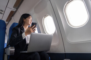 Traveling and technology. Flying at first class. Pretty young Asian business woman using smartphone and laptop while sitting in airplane