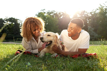 african american happy couple lie together with dog in park in summer, man and woman hug and love retriever