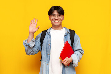 young guy asian student in denim shirt and glasses with backpack holding books and waving hello