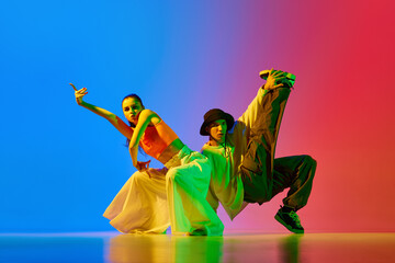 Talented, artistic, expressive young man and woman in modern clothes dancing hip hop against blue red background in neon. Concept of hobby, action, street style, contemporary dance, youth, fashion