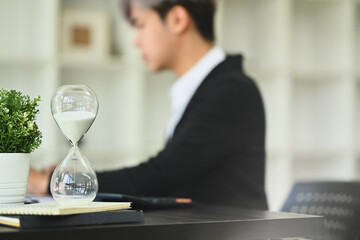 Hourglass on wooden office table with blurred businessman working on background. Time management...