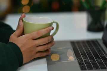Young woman in warm sweater holding cup of hot tea on background of Christmas tree with lights