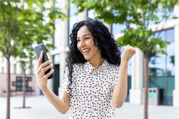 Young joyful woman winner received online notification on phone, Hispanic woman with curly hair...
