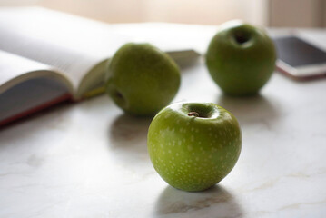 Green apples lie on a marble table. There is a telephone nearby. There is a phone and a book nearby