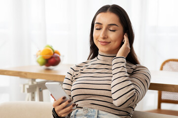 Cheerful pretty millennial arab lady with smartphone sit at sofa, in wireless headphones listen music
